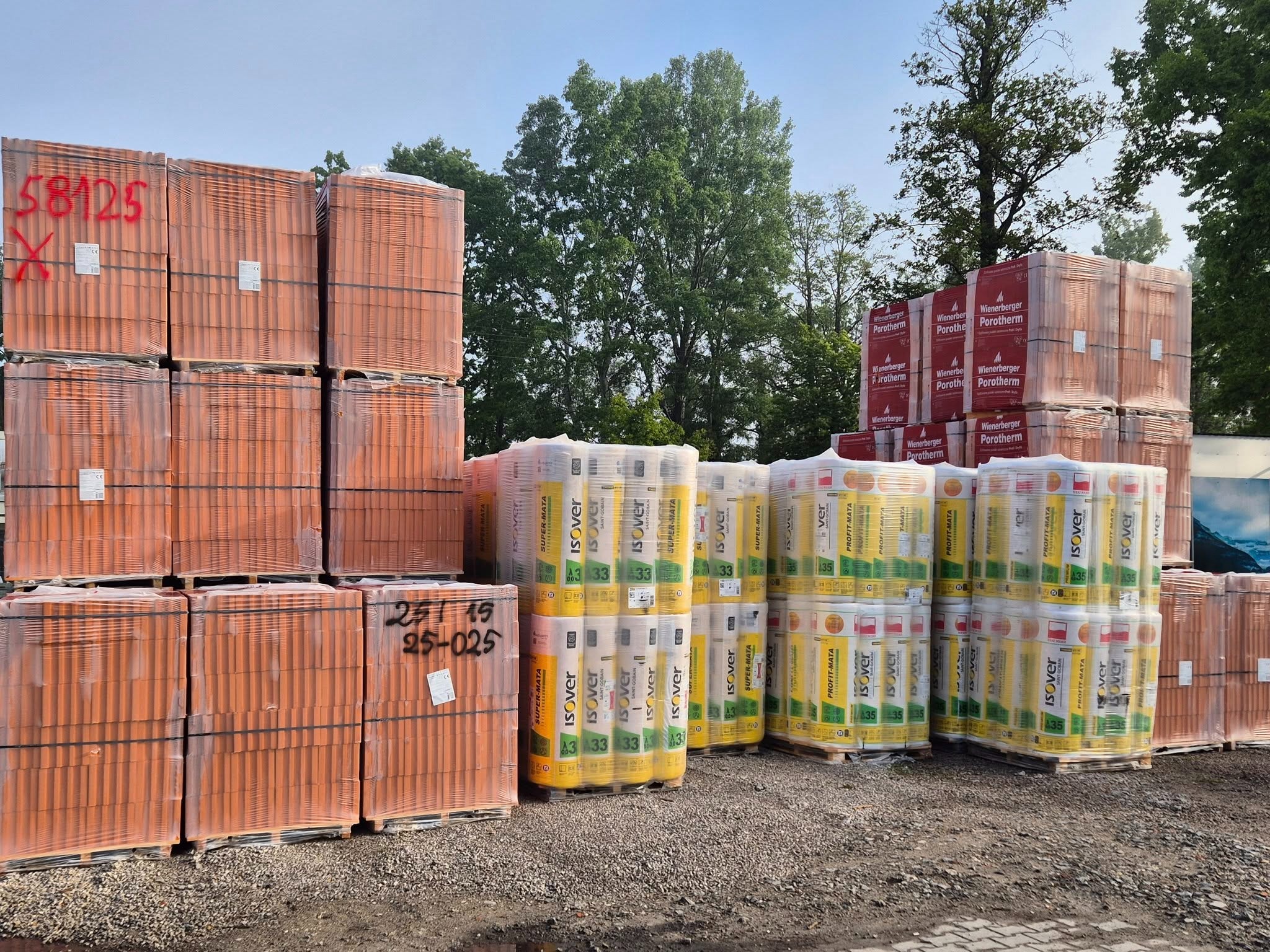 Składowane materiały budowlane: palety z czerwoną ceramiką Porotherm, żółtą wełną Isover, na szarym podłożu. Część materiałów oznaczona czerwoną farbą.