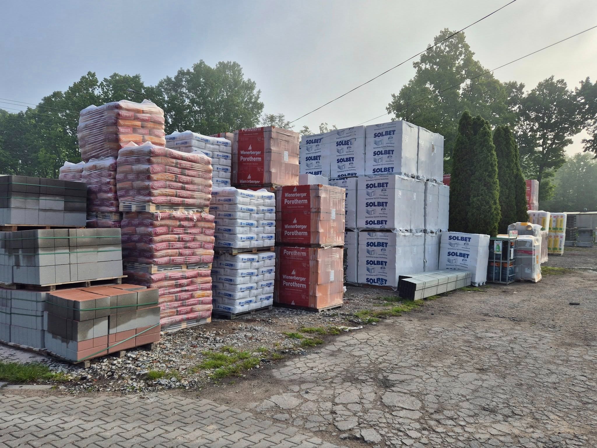 Składowisko materiałów budowlanych: palety z bloczkami Solbet, Wienerberger Porotherm i kostką brukową na zewnątrz, w tle zieleń drzew.