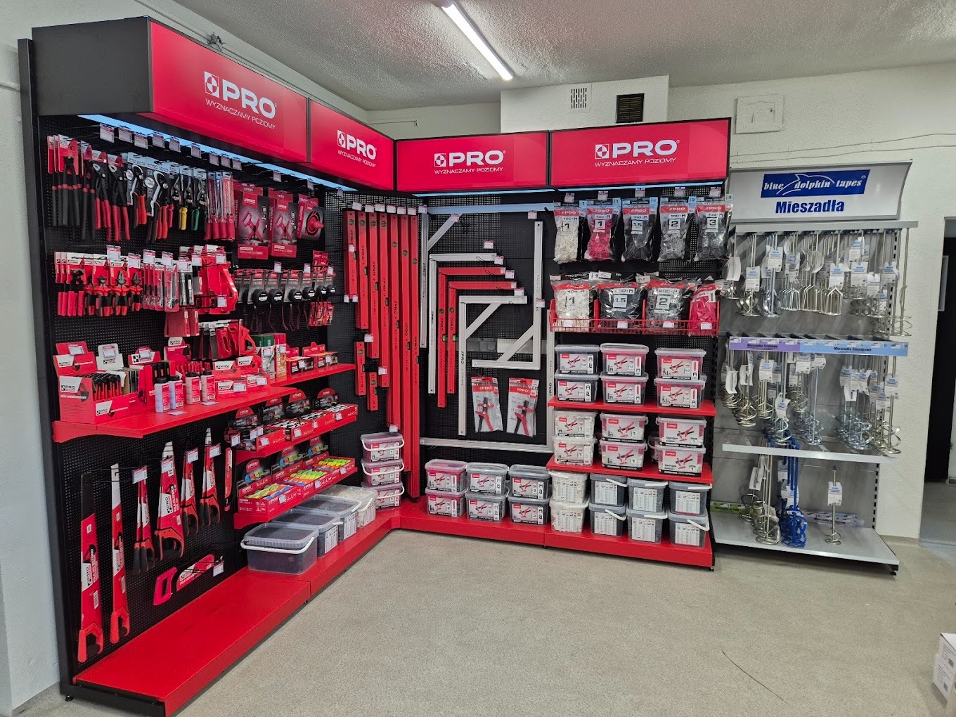 Ekspozycja narzędzi PRO w sklepie: poziomice, mieszadła, pędzle i inne akcesoria budowlane. Czerwono-czarne regały z logo PRO. Widok z perspektywy klienta.