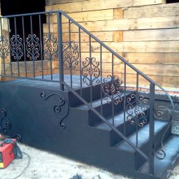 Rupol-Metal - Metalowe schody zewnętrzne z ozdobnymi poręczami, czarne, z widocznym sprzętem spawalniczym. Tło z desek i cegieł. Ujęcie z dołu, podkreślające detale.