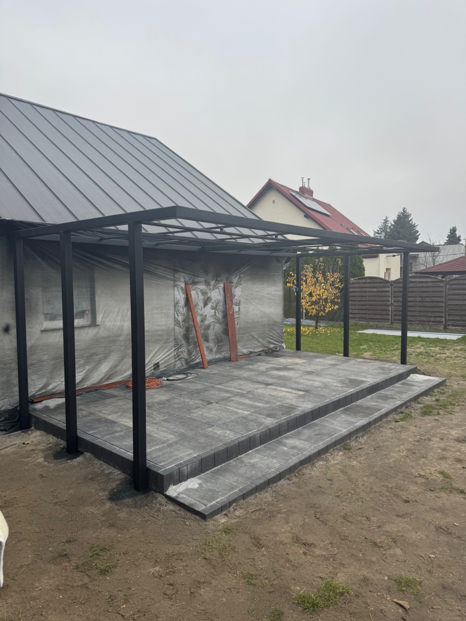Nowoczesna, czarna pergola metalowa zadaszająca taras z szarych płyt, z dwoma stopniami. Dom w tle częściowo zakryty folią. Pochmurny dzień.