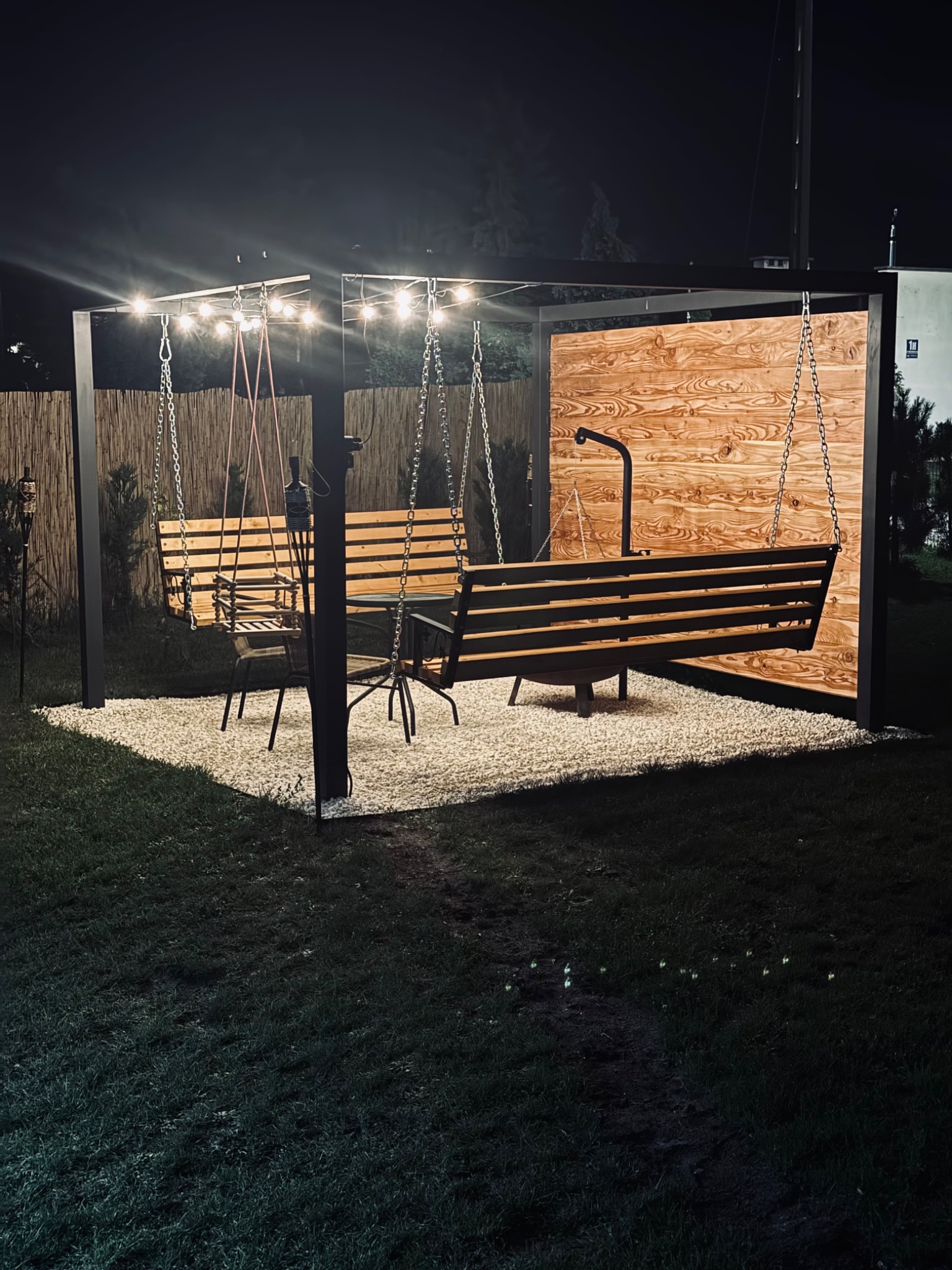 Oświetlona pergola z huśtawkami na łańcuchach, drewnianym panelem i kamiennym podłożem, otoczona zielonym trawnikiem w nocnej scenerii.