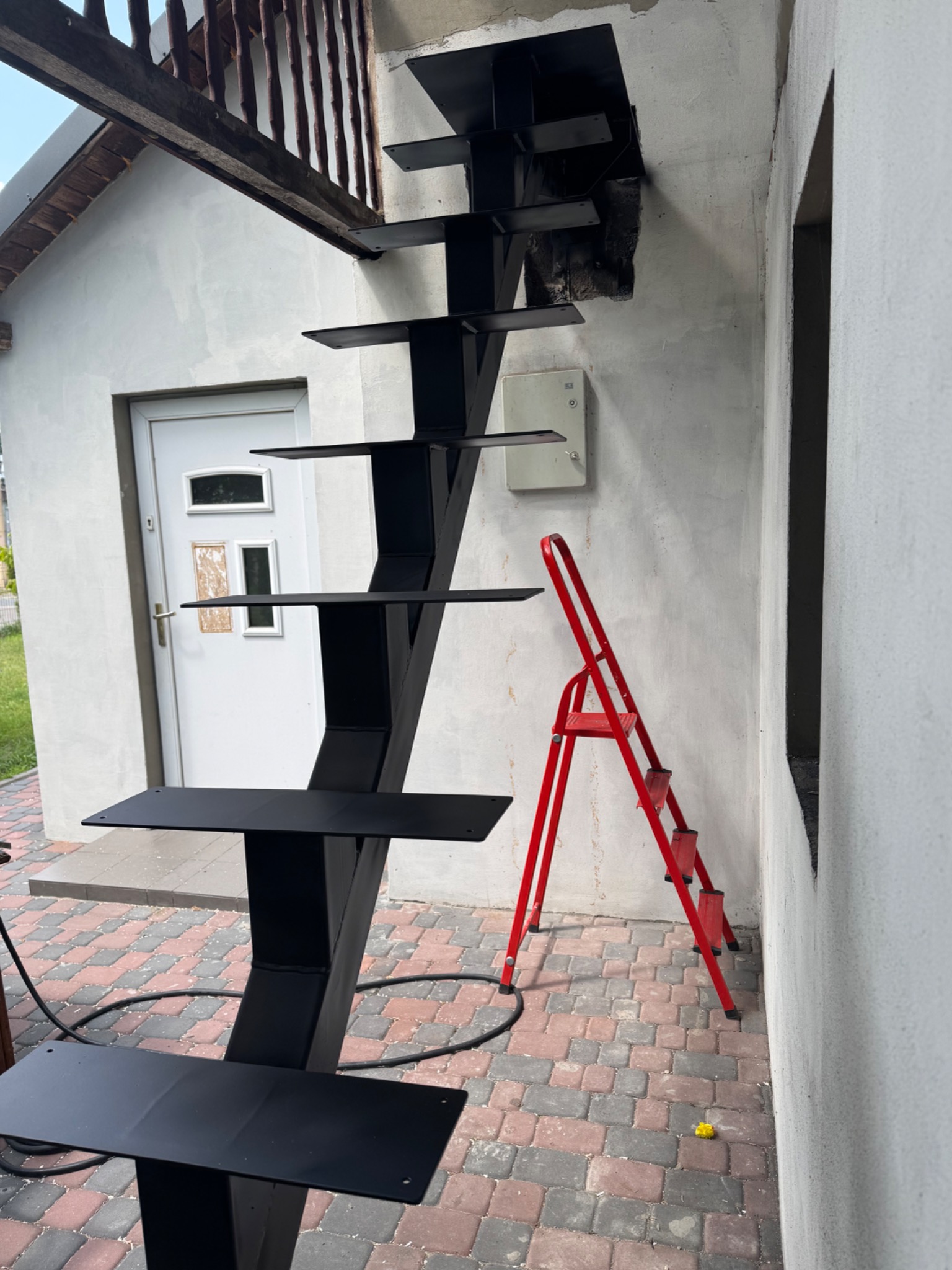 Nowoczesne, czarne schody metalowe zewnętrzne, z asymetryczną konstrukcją, prowadzące do otworu w ścianie budynku. Obok stoi czerwona drabina.