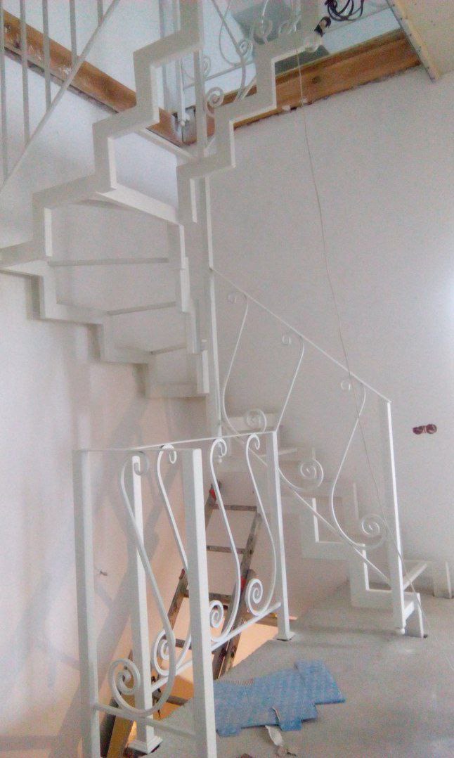 Białe, metalowe schody z ozdobnymi poręczami w jasnym wnętrzu. Widoczna drabina i fragment drewnianej konstrukcji stropu. Ujęcie z dołu, podkreślające spiralny kształt.