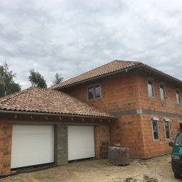 Usługi budowlane Franciszek Kulig - Budowa domu z garażem w Limanowej. Czerwona cegła, dachówka ceramiczna, białe bramy garażowe. Widoczny samochód dostawczy i materiały budowlane. Stan surowy otwarty.