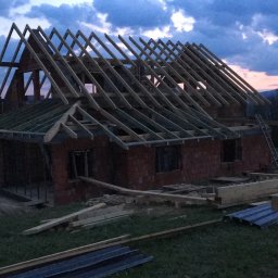 Usługi budowlane Franciszek Kulig - Budowa domu z czerwonej cegły w trakcie realizacji, widoczne drewniane elementy konstrukcji dachu na tle zachodzącego słońca i gór. Na pierwszym planie materiały budowlane.
