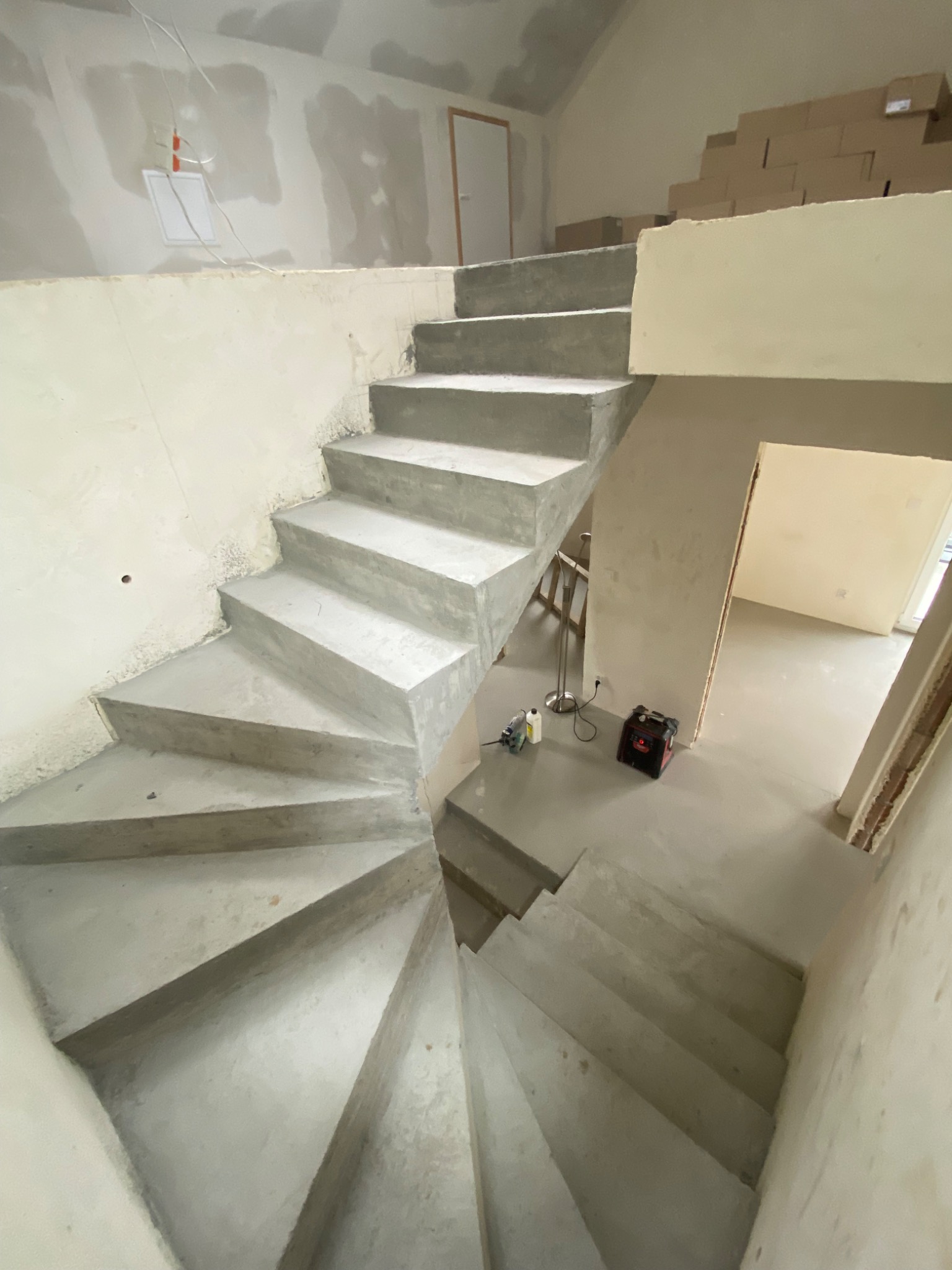 Surowe, betonowe schody spiralne w trakcie budowy, z widocznymi narzędziami i kartonami w tle, ukazujące etap prac wykończeniowych wewnątrz budynku.