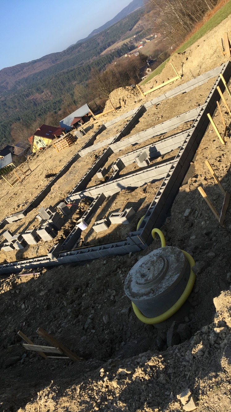 Fundamenty z bloczków betonowych na nierównym terenie, widok z góry, z elementami instalacji. W tle góry i zabudowania.