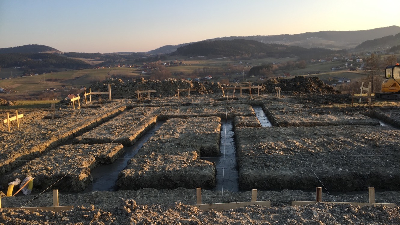Wykopane i częściowo zalane fundamenty domu jednorodzinnego na tle pagórkowatego krajobrazu oświetlonego wschodzącym słońcem. Widoczne drewniane szalunki i wytyczone sznurki.