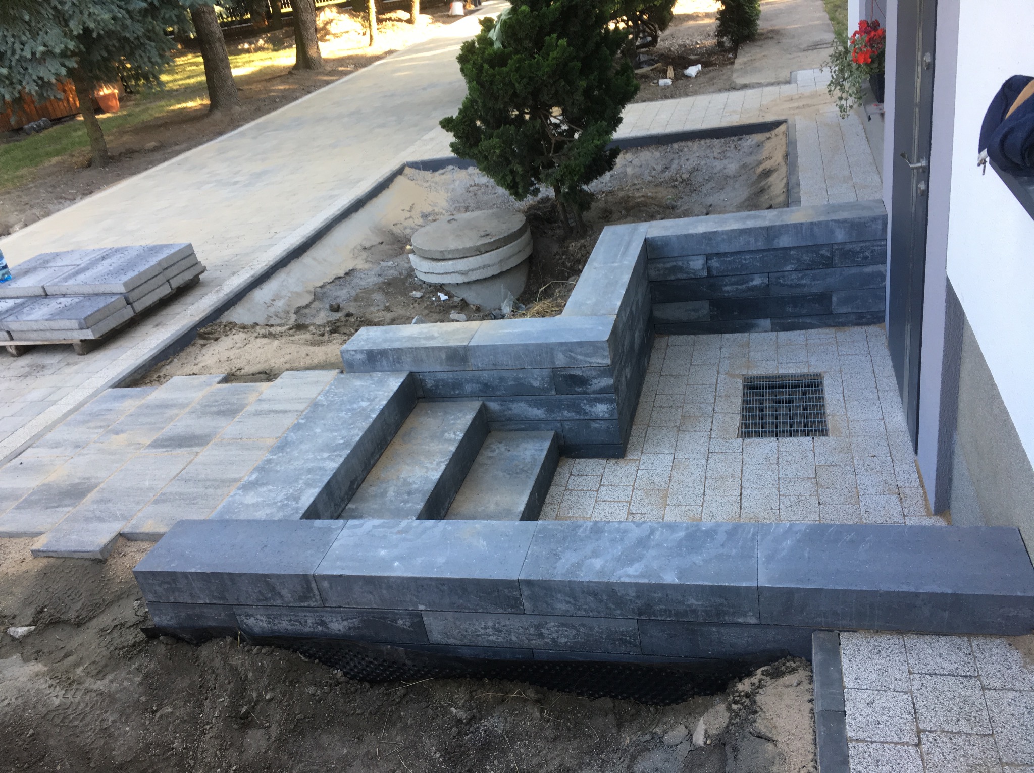 Układanie kostki brukowej na posesji: szara kostka, obrzeża z bloków, studzienka kanalizacyjna, widoczne warstwy podbudowy. Prace w toku, teren częściowo niezagospodarowany.