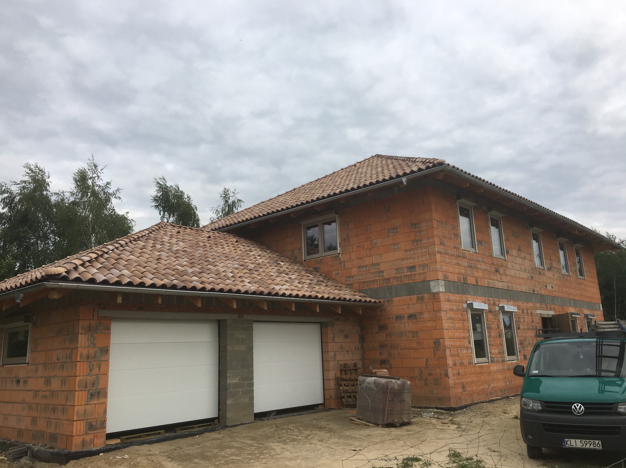 Budowa domu z garażem w Limanowej. Czerwona cegła, dachówka ceramiczna, białe bramy garażowe. Widoczny samochód dostawczy i materiały budowlane. Stan surowy otwarty.