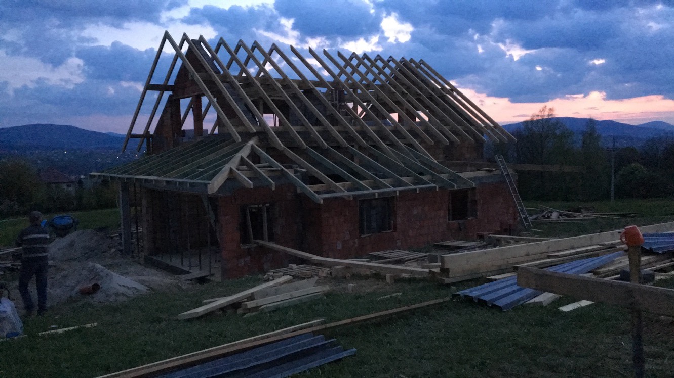 Budowa domu z czerwonej cegły w trakcie realizacji, widoczne drewniane elementy konstrukcji dachu na tle zachodzącego słońca i gór. Na pierwszym planie materiały budowlane.