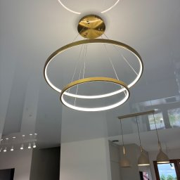 Firma Remontowo Budowlana LOFTIS Sebastian Jakubowski - Błyszczący sufit napinany z nowoczesnym, złotym oświetleniem LED w kształcie okręgów. Widoczne inne lampy w tle. Minimalistyczny design wnętrza.