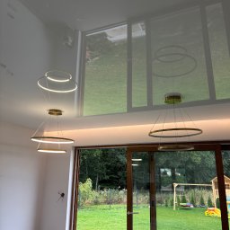 Firma Remontowo Budowlana LOFTIS Sebastian Jakubowski - Nowoczesny, okrągły żyrandol z LED, zawieszony w jasnym pokoju z dużymi oknami wychodzącymi na zielony ogród. Widoczne odbicie żyrandola w błyszczącej powierzchni sufitu.