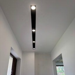 Firma Remontowo Budowlana LOFTIS Sebastian Jakubowski - Białe wnętrze z nowoczesnym oświetleniem sufitowym: czarny, podłużny panel z wbudowanymi, okrągłymi lampami LED. Widoczne drzwi i okno w tle.