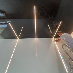 Firma Remontowo Budowlana LOFTIS Sebastian Jakubowski - Instalacja oświetlenia liniowego LED na styku ściany i sufitu przez montera na drabinie. Widoczne rękawice ochronne i precyzyjne łączenie elementów.