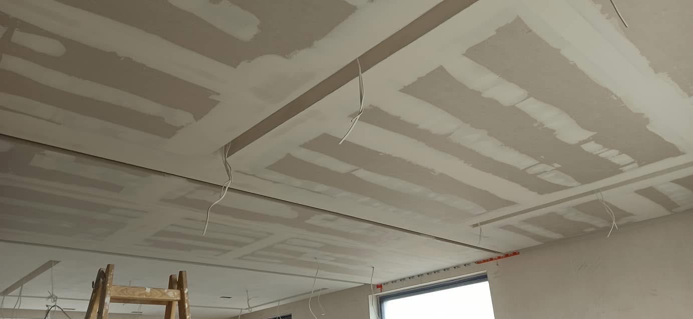 Sufit z płyt gipsowo-kartonowych w trakcie szpachlowania, widoczne kable i drabina. Nierównomiernie nałożona gładź, przygotowanie do dalszych prac wykończeniowych.