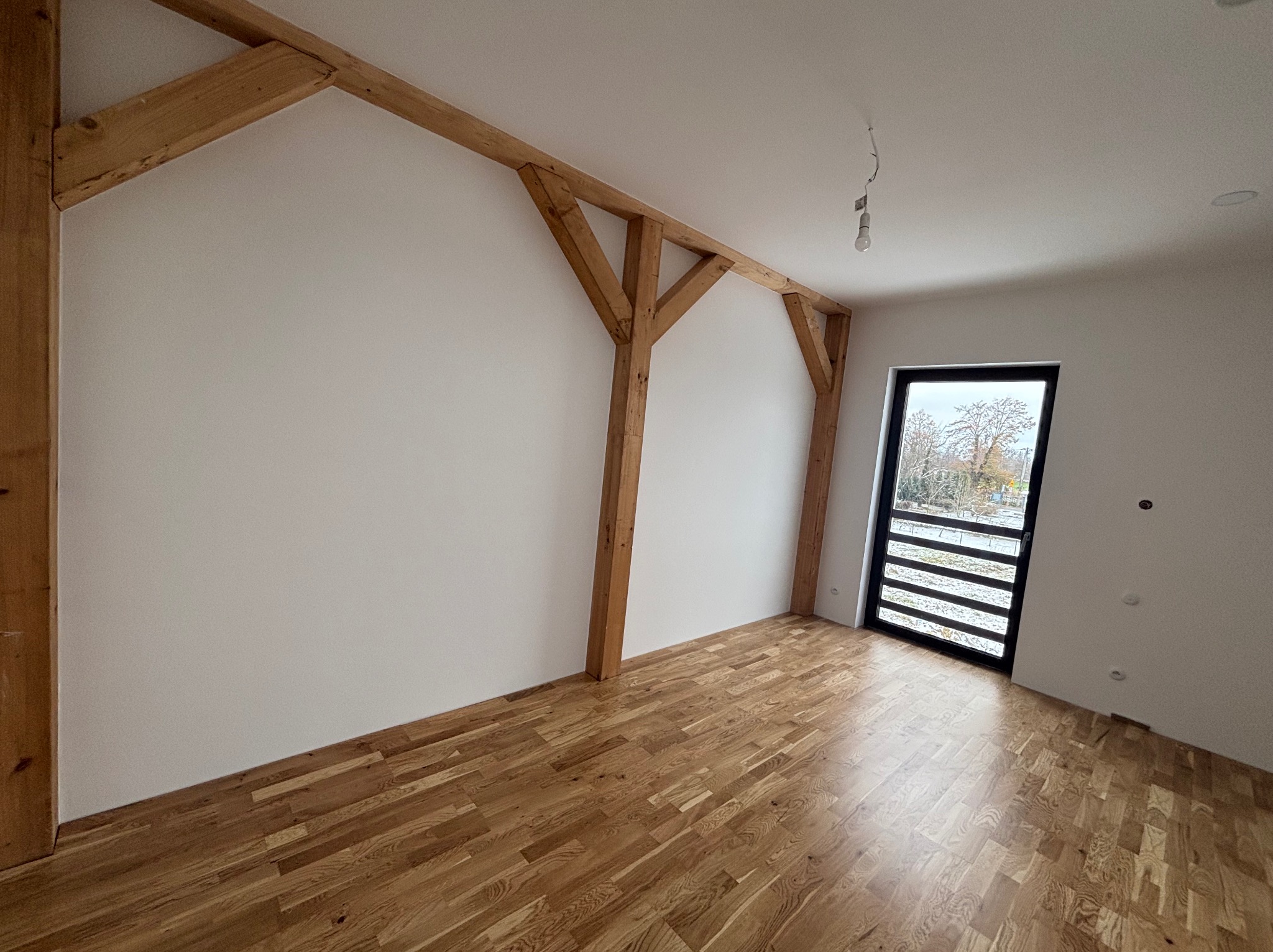 Jasne wnętrze z drewnianą podłogą i elementami konstrukcyjnymi z drewna. Widoczne białe ściany i czarne ramy okienne. Surowy stan wykończenia z wiszącą żarówką.