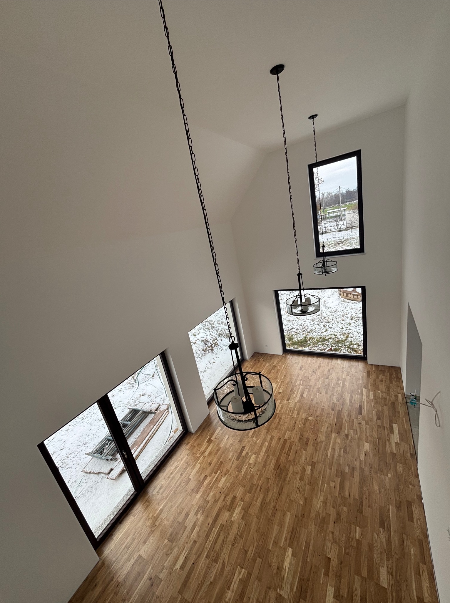 Wysoki, jasny pokój z parkietem i trzema oknami z widokiem na zimowy krajobraz. Dwie czarne, wiszące lampy dodają nowoczesnego charakteru wnętrzu.