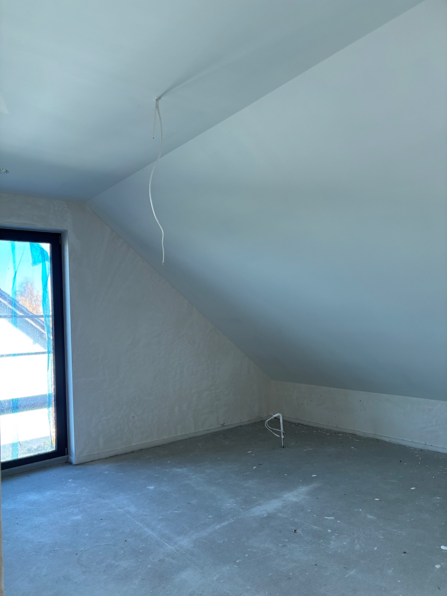 Pokój na poddaszu po szpachlowaniu ścian i sufitu, widoczne gładkie powierzchnie, okno z folią ochronną i wystające przewody. Surowy stan wykończenia.