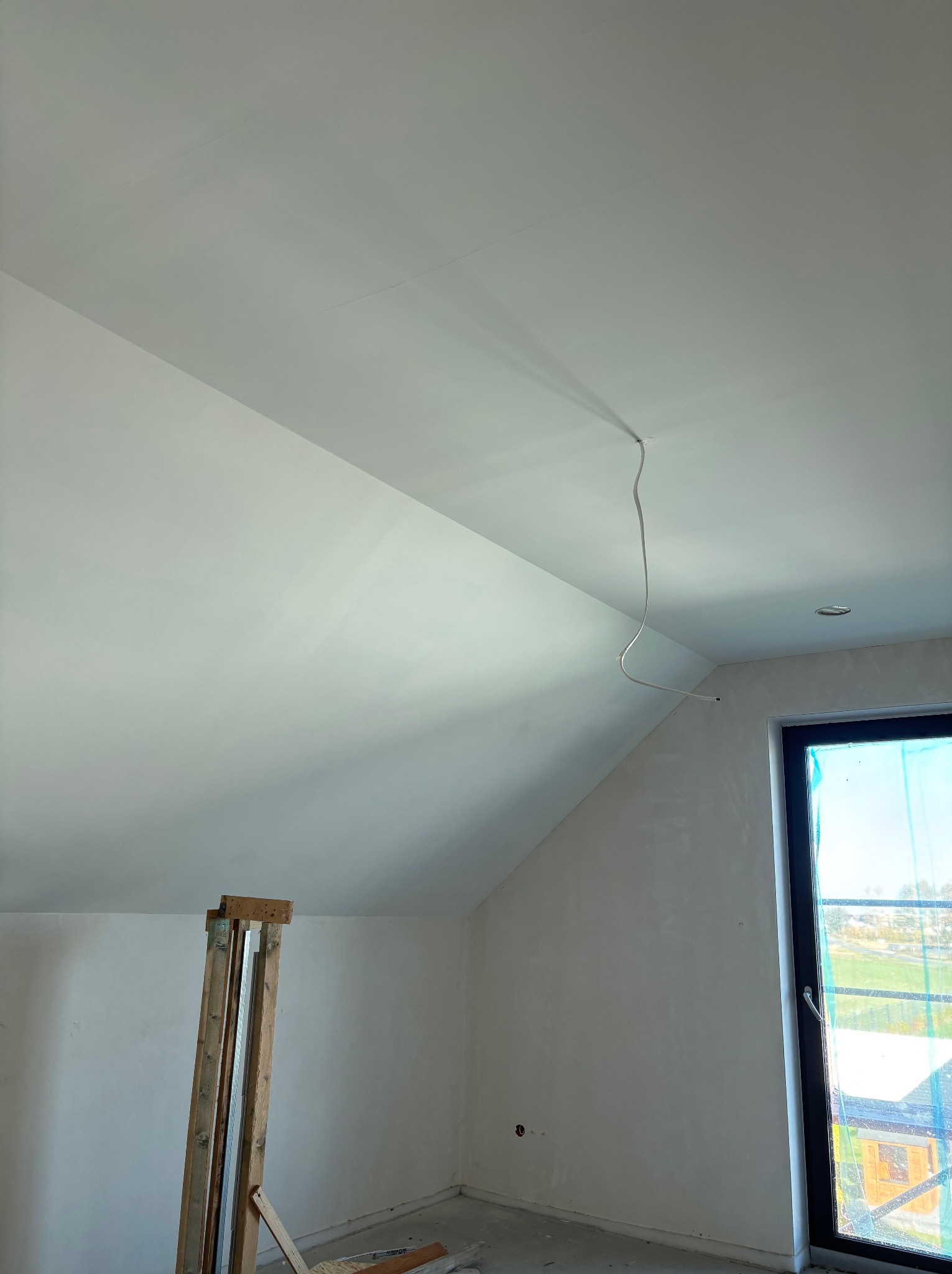 Pokój na poddaszu w trakcie prac wykończeniowych. Widoczne ściany po szpachlowaniu, okno z czarną ramą i wystający kabel z sufitu. Na podłodze resztki materiałów budowlanych.