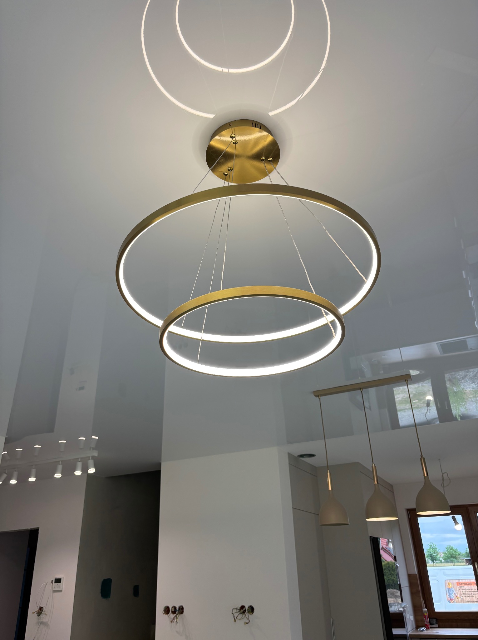 Błyszczący sufit napinany z nowoczesnym, złotym oświetleniem LED w kształcie okręgów. Widoczne inne lampy w tle. Minimalistyczny design wnętrza.