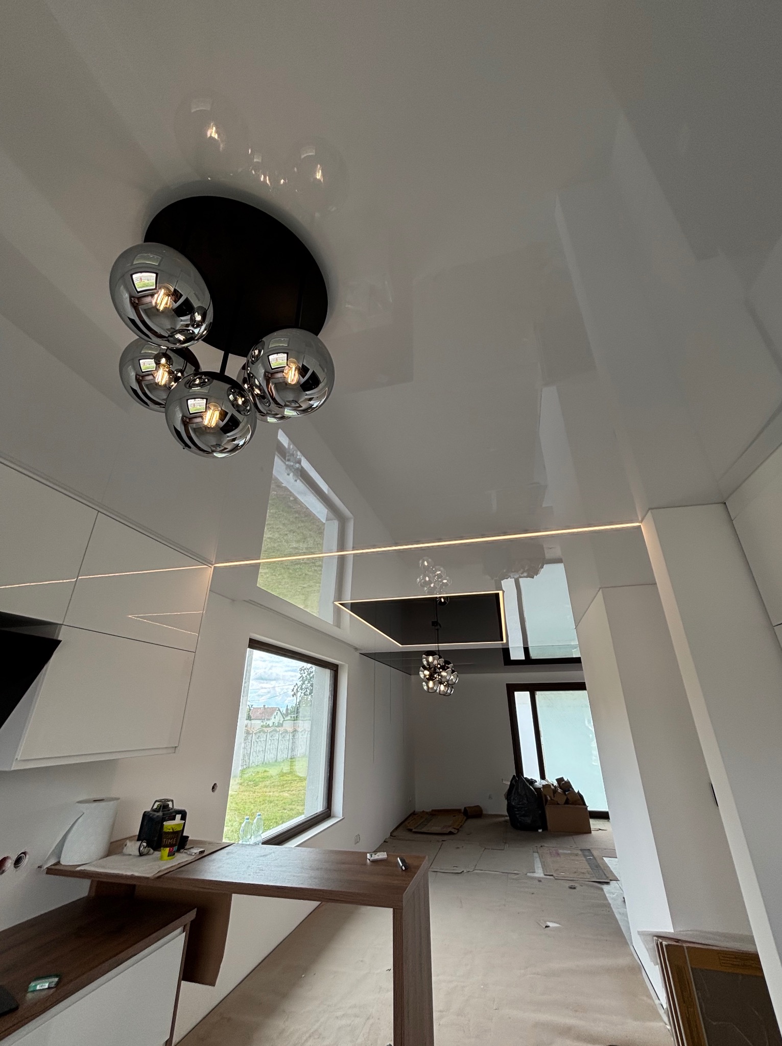 Biały, błyszczący sufit napinany z nowoczesnym oświetleniem kulistym w przestronnym wnętrzu. Widoczne okna i elementy wykończeniowe. Minimalistyczny design.