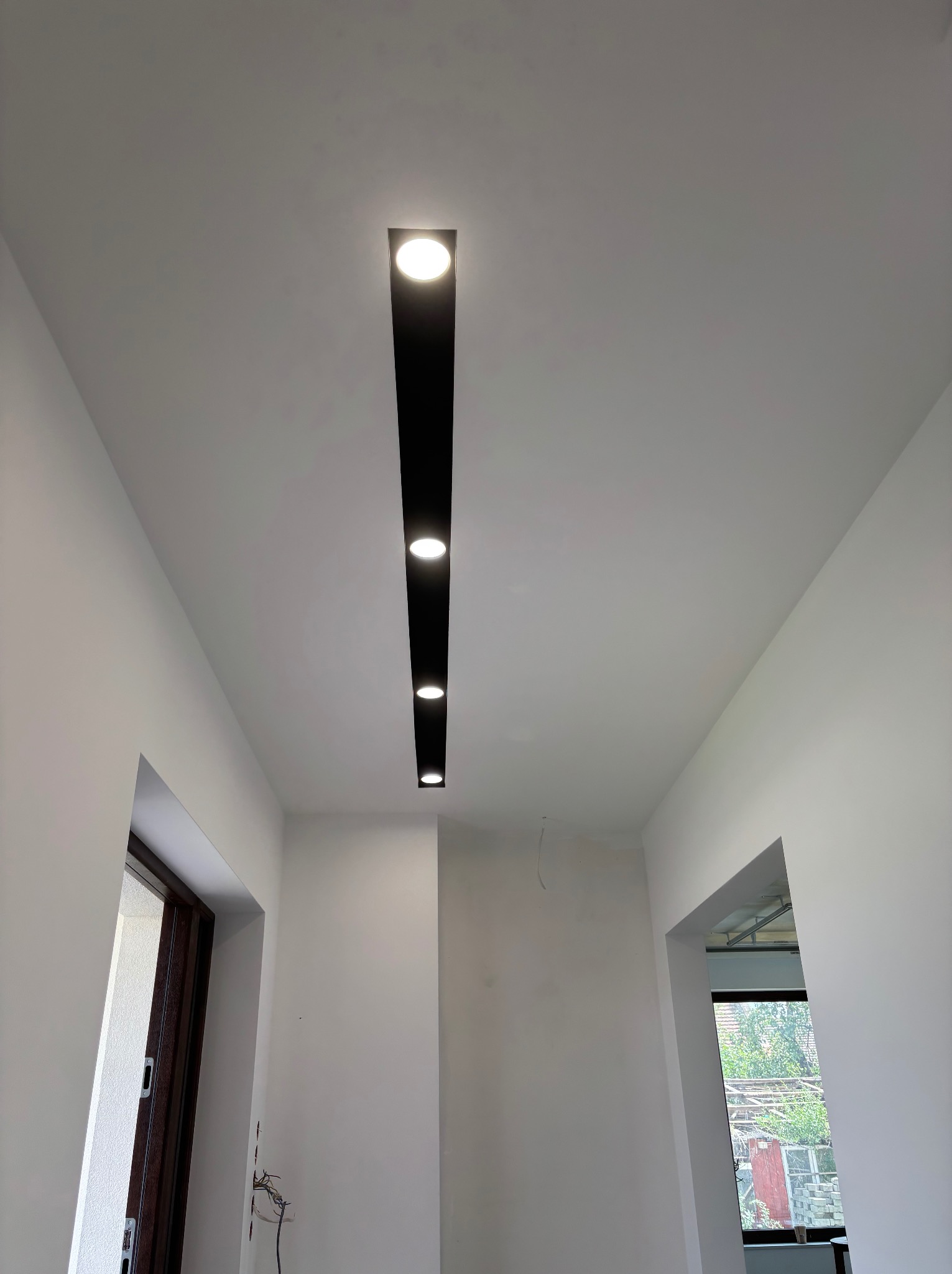 Białe wnętrze z nowoczesnym oświetleniem sufitowym: czarny, podłużny panel z wbudowanymi, okrągłymi lampami LED. Widoczne drzwi i okno w tle.