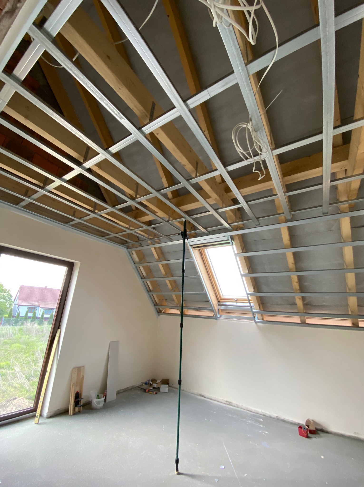 Stelaż sufitowy z profili metalowych w poddaszu, widok z dołu, okno dachowe, narzędzia i materiały budowlane na podłodze, przygotowanie do dalszych prac wykończeniowych.