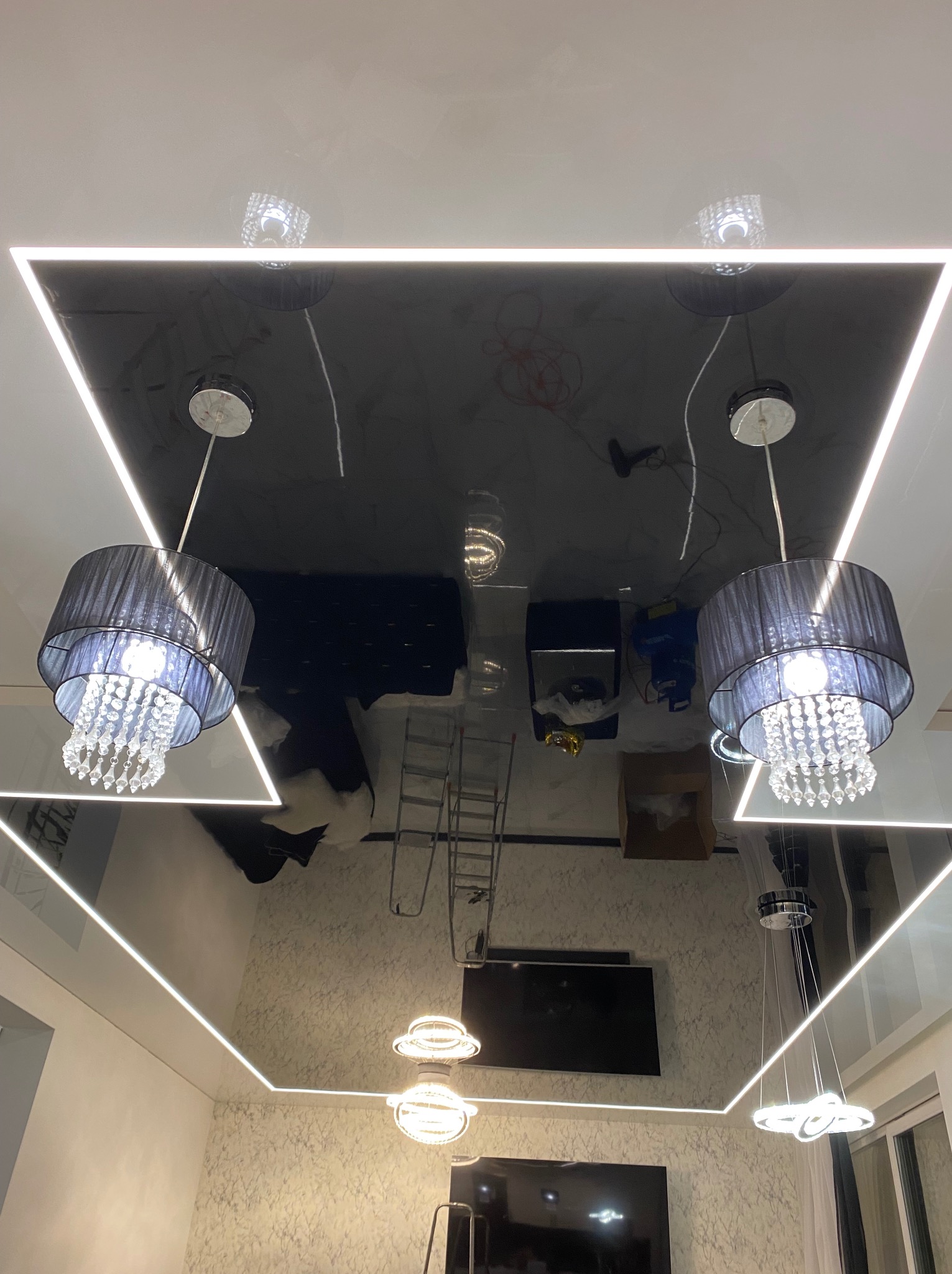 Czarny, błyszczący sufit napinany z oświetleniem LED w kształcie kwadratu i dwoma wiszącymi lampami z kryształkami, odbijający wnętrze pokoju.