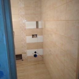 Jarosław Hekert - Beżowe płytki w łazience, częściowo ułożone, widoczne instalacje. Na ścianie biało-beżowy pas dekoracyjny. Podłoga wyłożona płytkami imitującymi drewno.