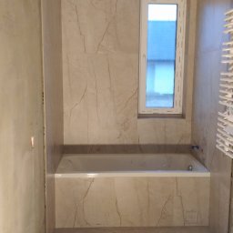 Jarosław Hekert - Wykończona łazienka z wanną w zabudowie, marmurowe płytki na ścianach, okno i grzejnik drabinkowy. Widoczne ślady po remoncie.