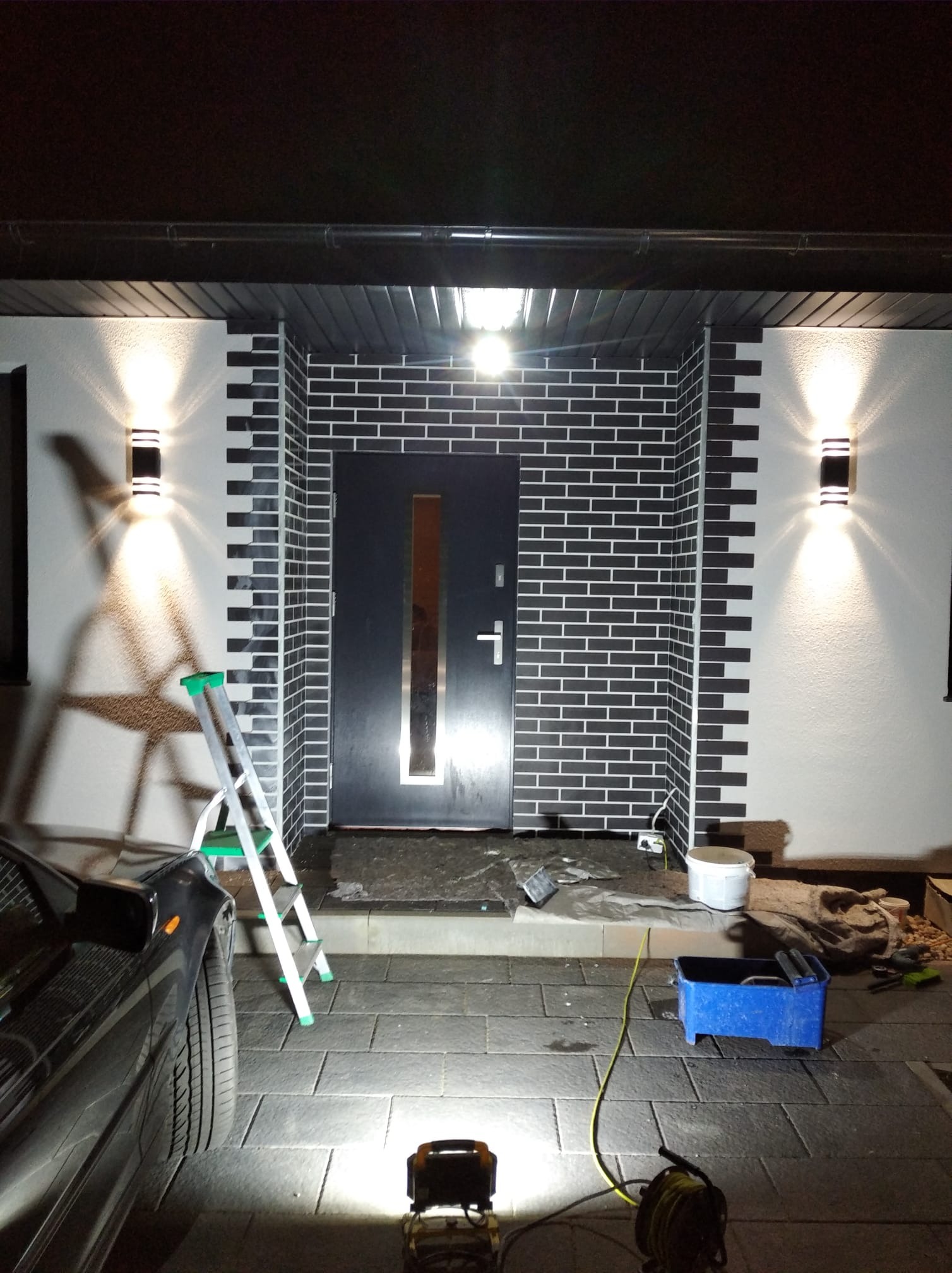 Wykończenie elewacji wejściowej z czarnej cegły klinkierowej z białą fugą, oświetlenie zewnętrzne, drabina i narzędzia na brukowanym podjeździe. Nocne ujęcie.