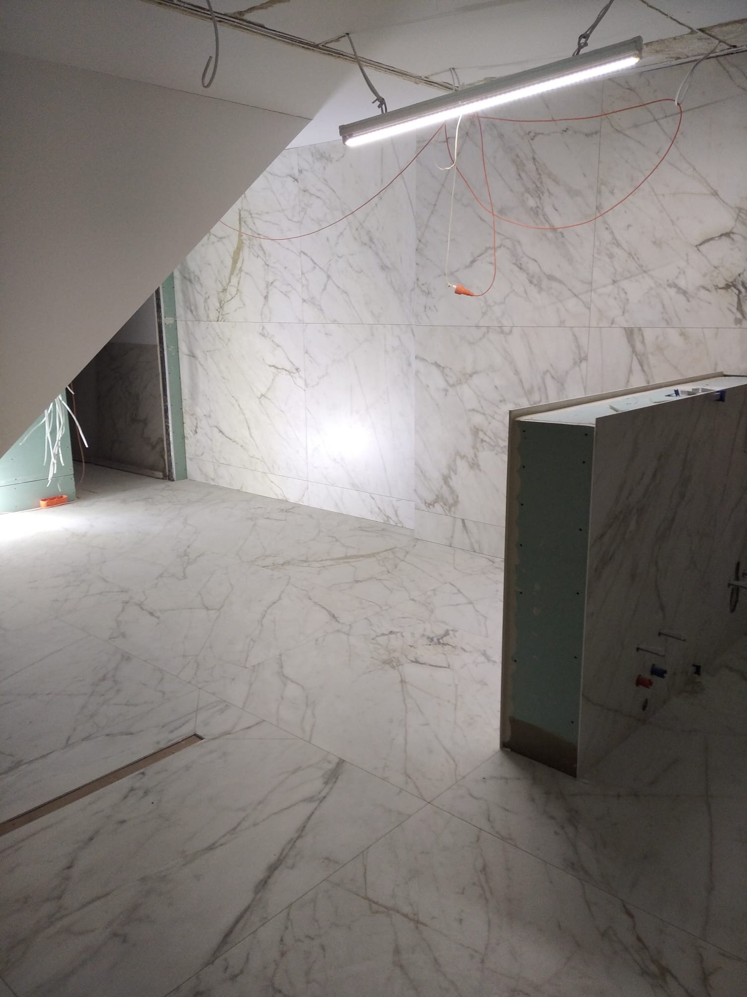 Wykończona łazienka z marmurowymi płytkami na ścianach i podłodze. Widoczna zabudowa z płyt gipsowo-kartonowych i instalacja elektryczna. Oświetlenie sufitowe.