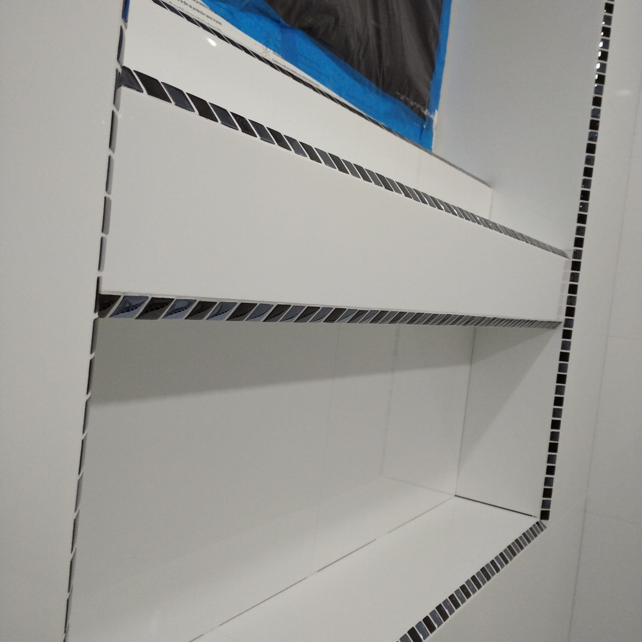 Biała półka wnękowa w łazience, wykończona czarną mozaiką. Widoczne zabezpieczenie folią malarską. Minimalistyczny design, precyzyjne ułożenie płytek.