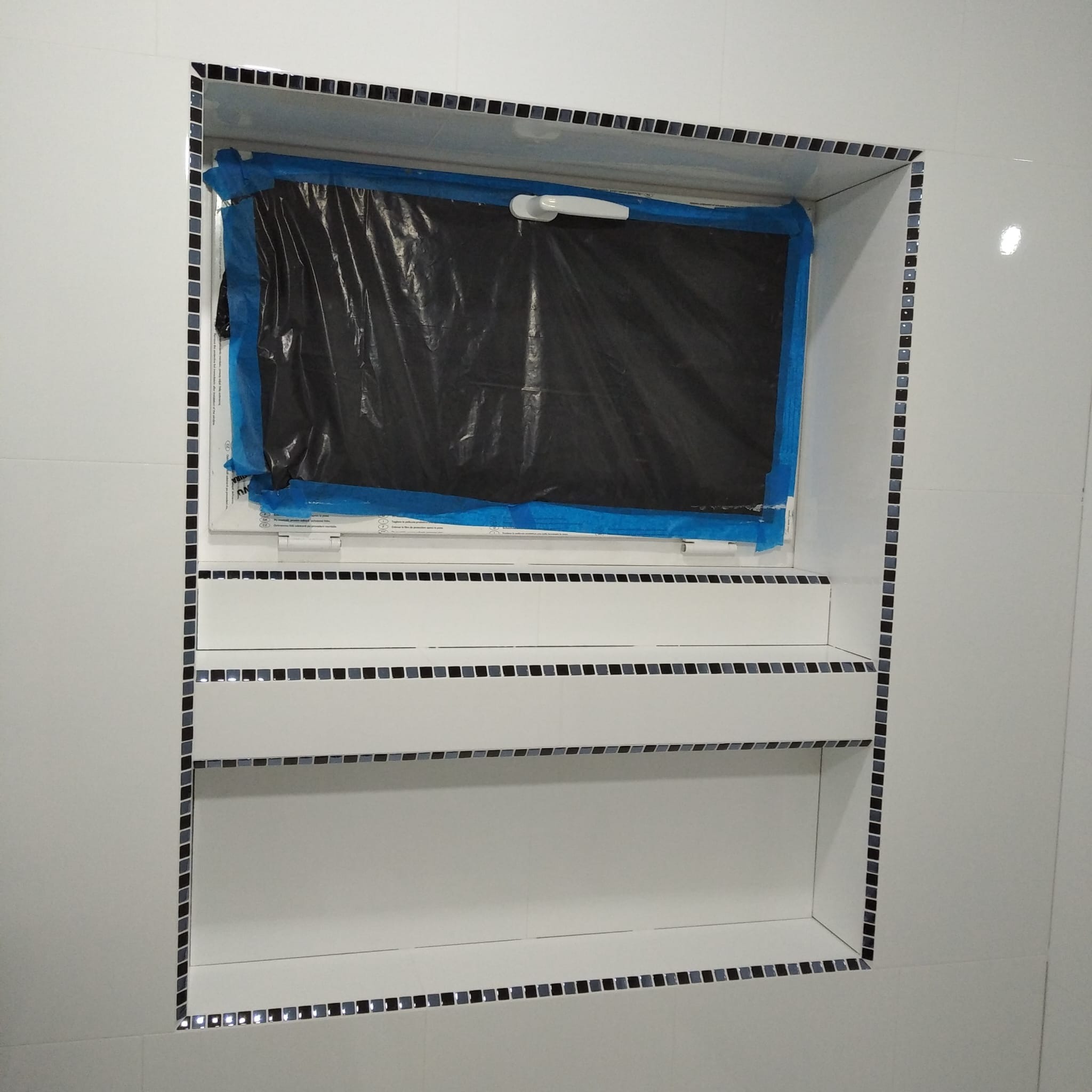 Narożna wnęka w łazience wykończona białą glazurą i mozaiką w odcieniach szarości i czerni. Okno zabezpieczone folią malarską. Widok z bliska, oświetlenie naturalne.