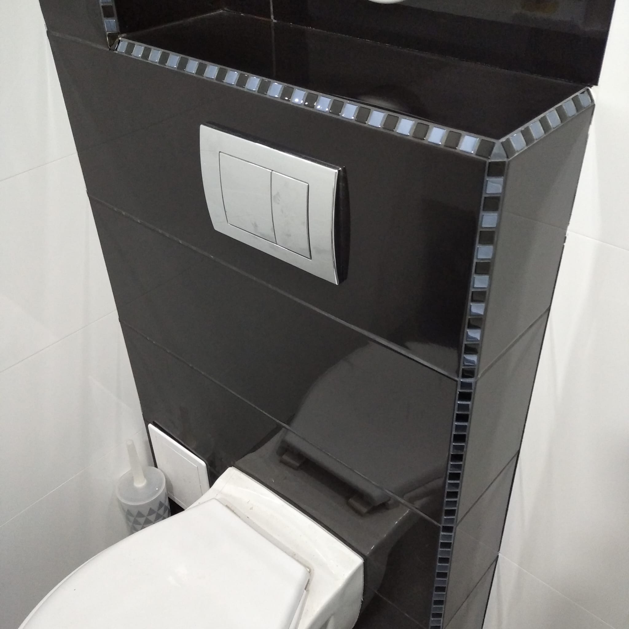 Czarna, błyszcząca zabudowa WC z mozaiką na krawędziach, widoczny przycisk spłukiwania i fragment toalety. Białe płytki w tle. Nowoczesna aranżacja łazienki.