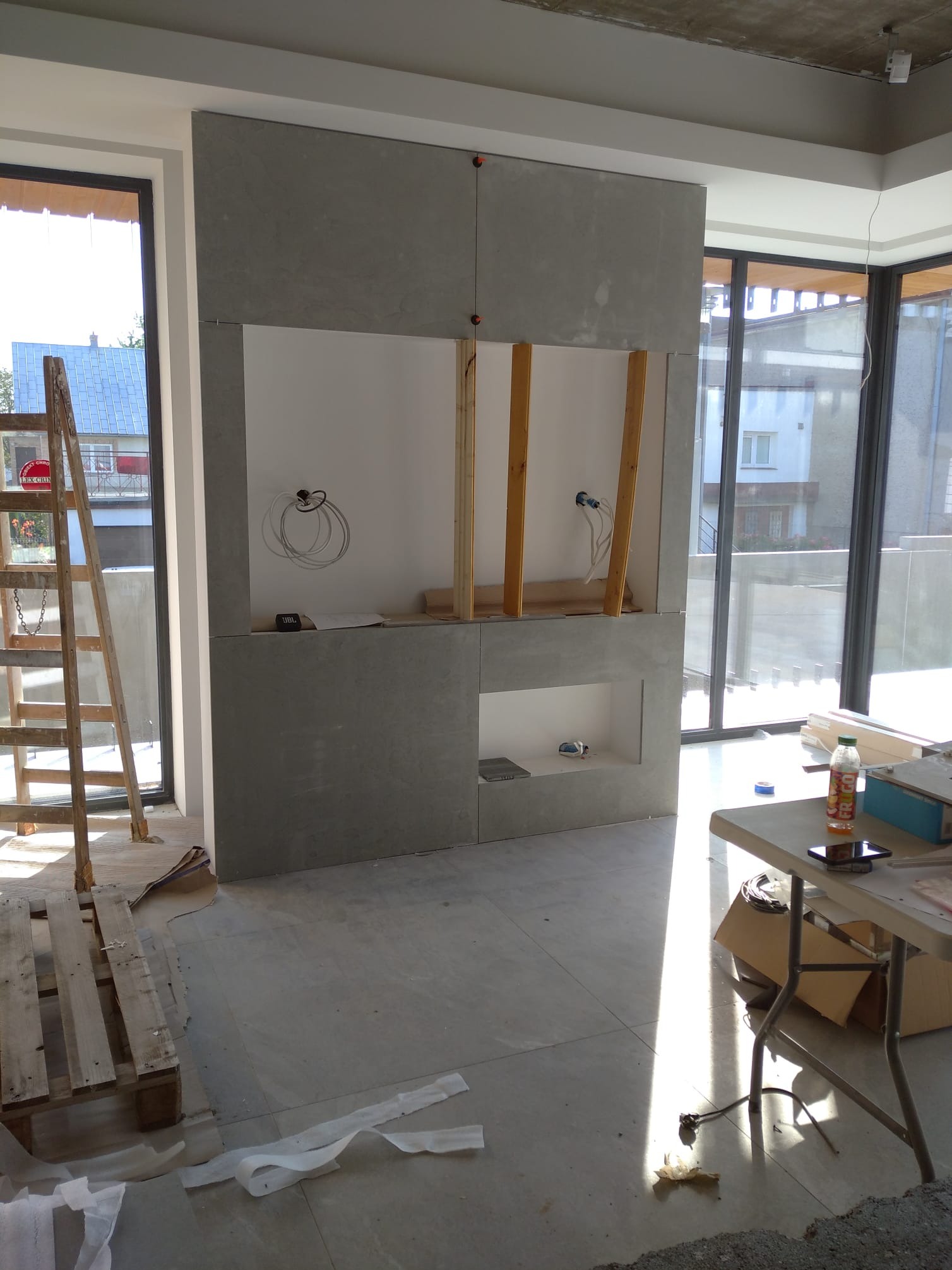 Wnętrze w trakcie prac wykończeniowych: ściana z szarej płyty z wnękami, widoczne instalacje elektryczne, drabina i stół z narzędziami. Duże okna w tle.
