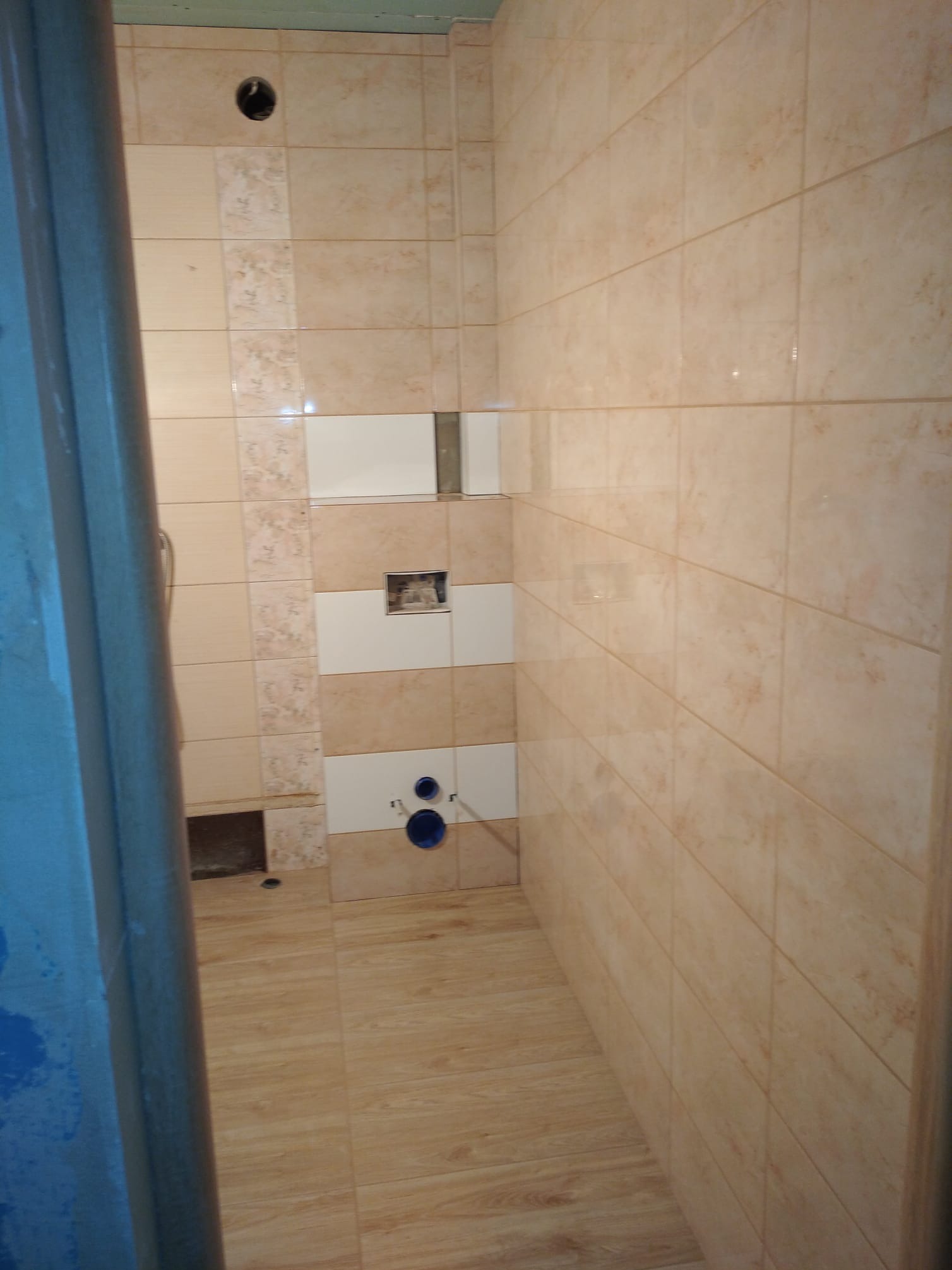 Beżowe płytki w łazience, częściowo ułożone, widoczne instalacje. Na ścianie biało-beżowy pas dekoracyjny. Podłoga wyłożona płytkami imitującymi drewno.