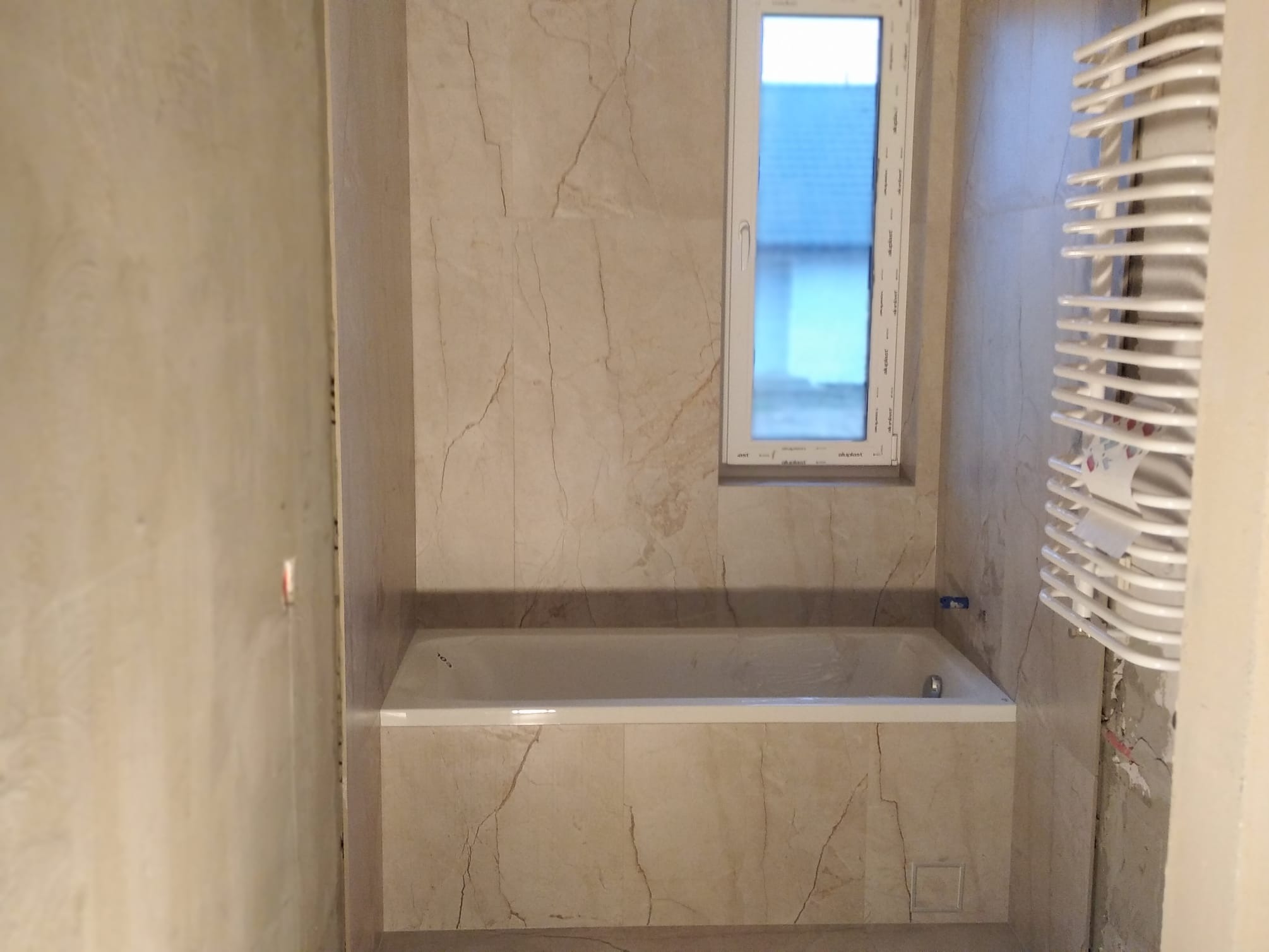 Wykończona łazienka z wanną w zabudowie, marmurowe płytki na ścianach, okno i grzejnik drabinkowy. Widoczne ślady po remoncie.