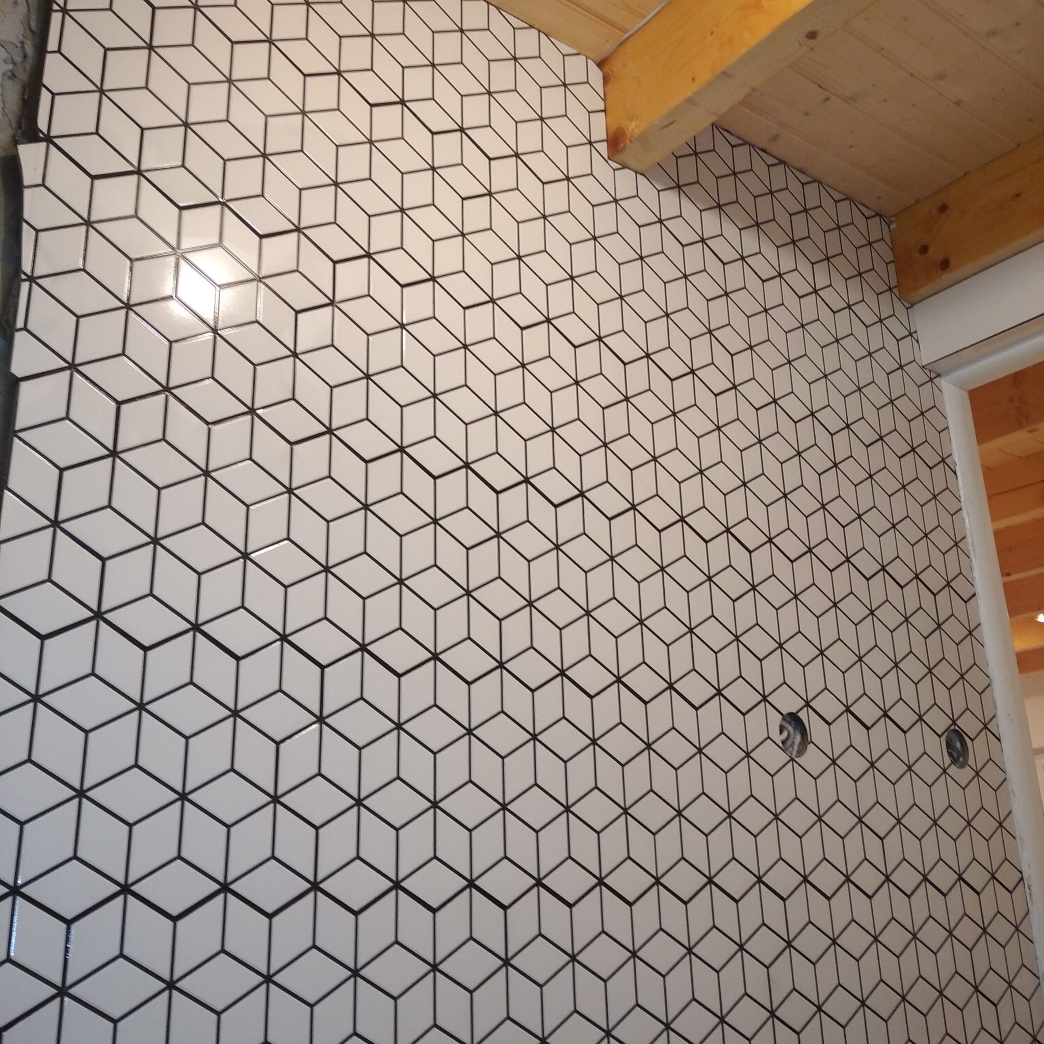 Ściana wyłożona białymi, sześciennymi płytkami z czarną fugą, tworzącymi iluzję 3D. Widoczne drewniane elementy konstrukcyjne sufitu i otwory na instalacje.
