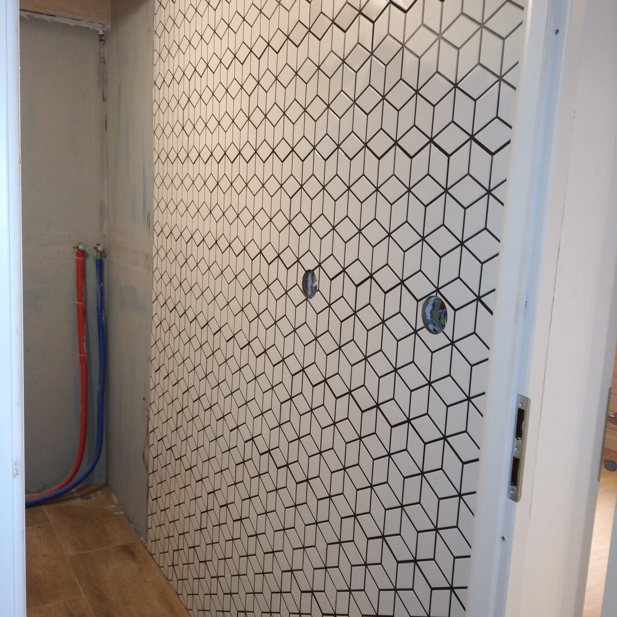 Ściana wyłożona białą mozaiką w geometryczny wzór 3D, z widocznymi otworami na instalacje. Fragment podłogi z płytek i rury instalacyjne w tle.