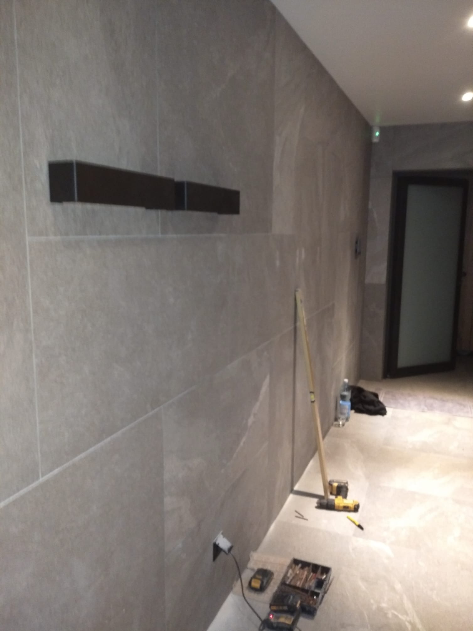 Wykończona ściana z szarych płytek w łazience, z zamontowanymi półkami. Na podłodze narzędzia glazurnicze i poziomica. Widoczny fragment drzwi.