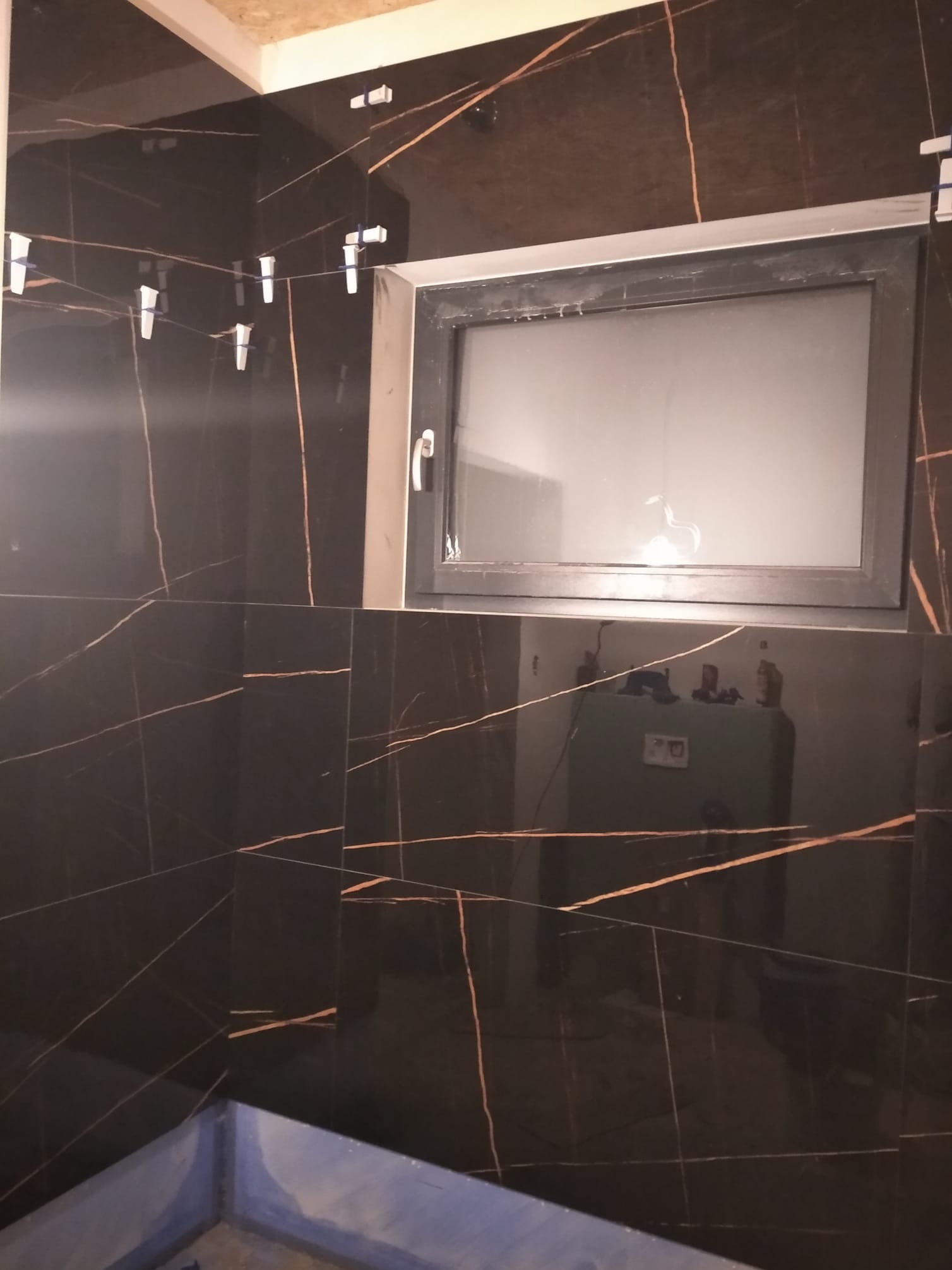 Częściowo wykończona łazienka z czarnymi płytkami imitującymi marmur, z widocznymi krzyżykami dystansowymi i oknem. W tle odbicie wyposażenia łazienki.