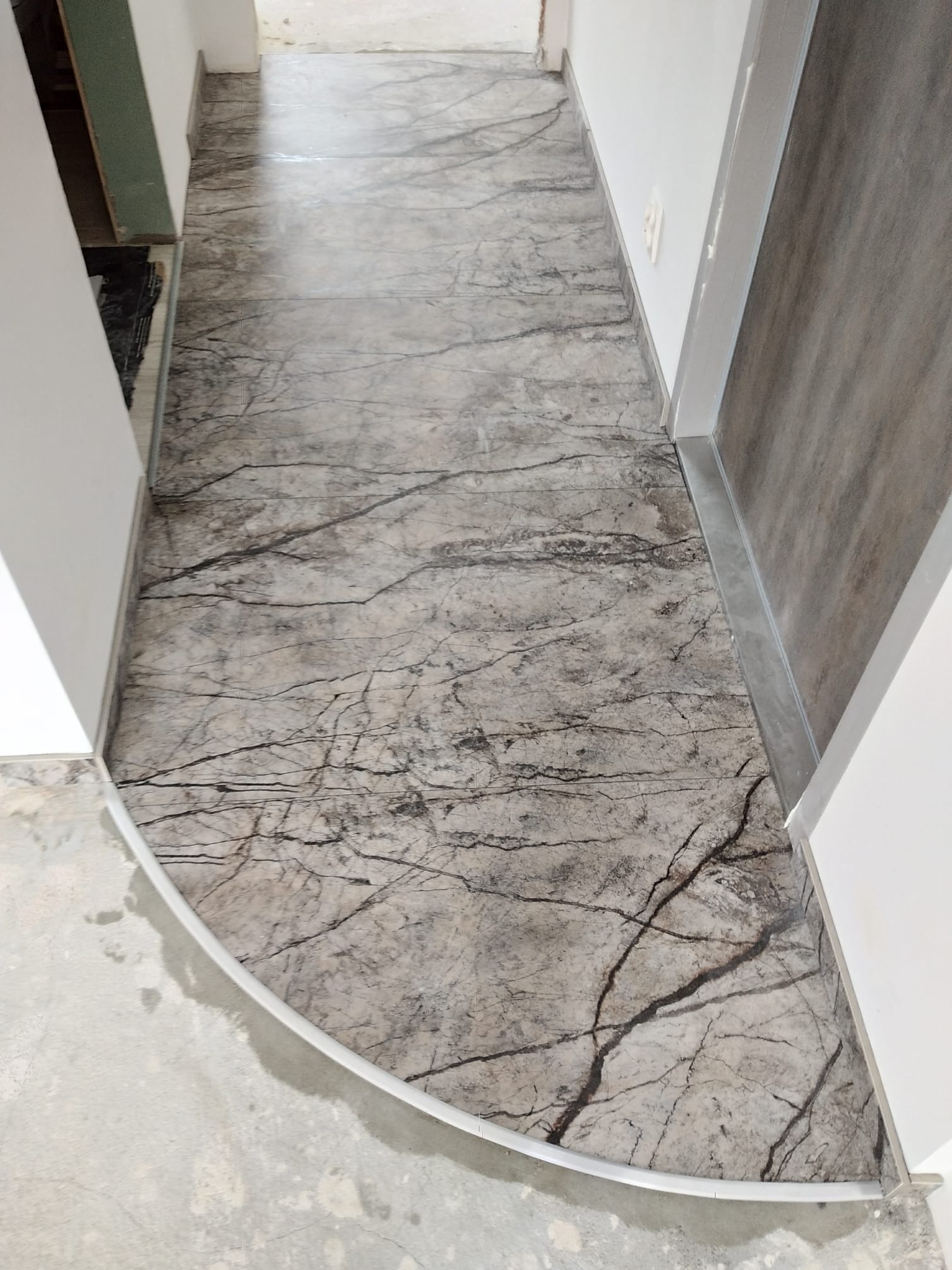 Ułożona podłoga z płytek imitujących marmur w korytarzu, z widocznymi listwami przypodłogowymi i fragmentem surowej posadzki przed wejściem do pomieszczenia.