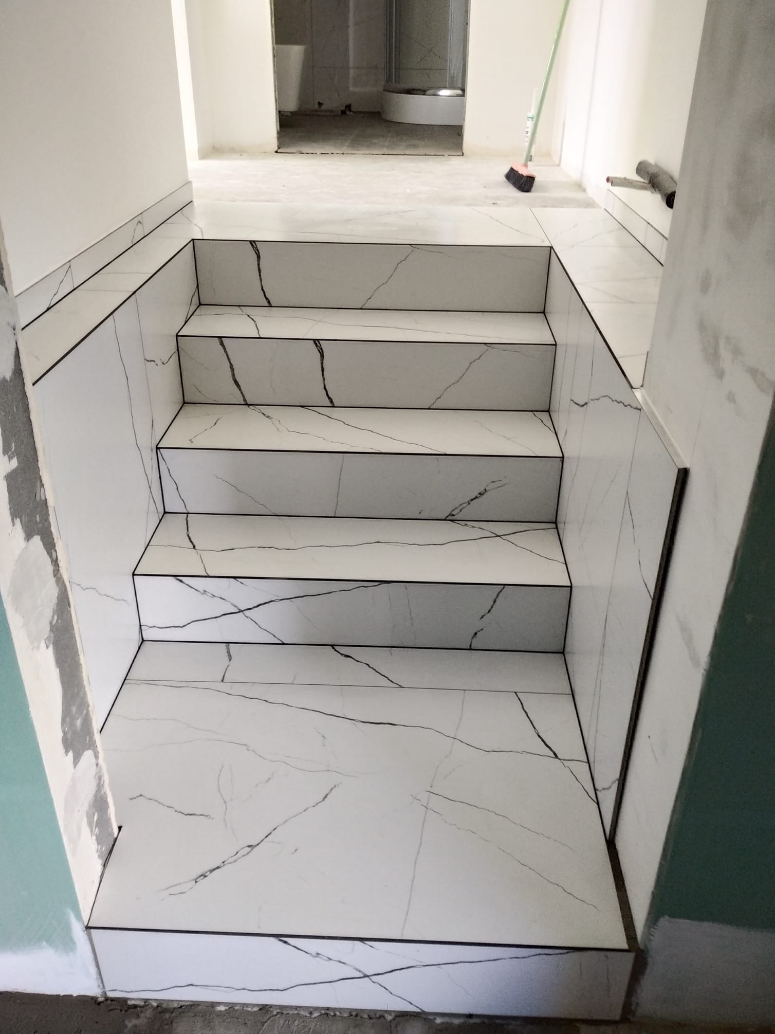 Schody wyłożone białymi płytkami imitującymi marmur z czarnymi żyłami, widok z góry, w tle fragment łazienki. Wykończenie schodów z dbałością o detale.