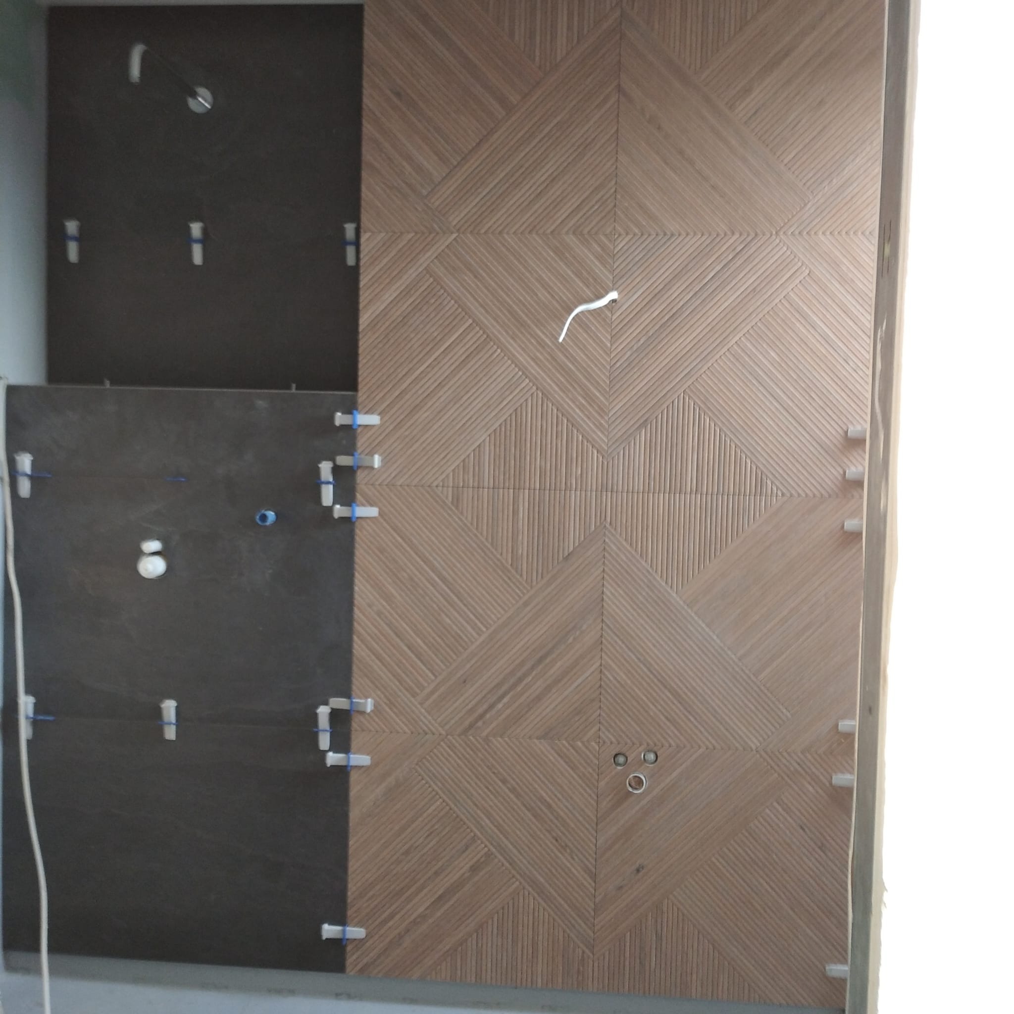 Układanie dekoracyjnej, drewnopodobnej glazury na ścianie z widocznymi krzyżykami dystansowymi i otworami na armaturę. Częściowo ułożona ciemna glazura obok.