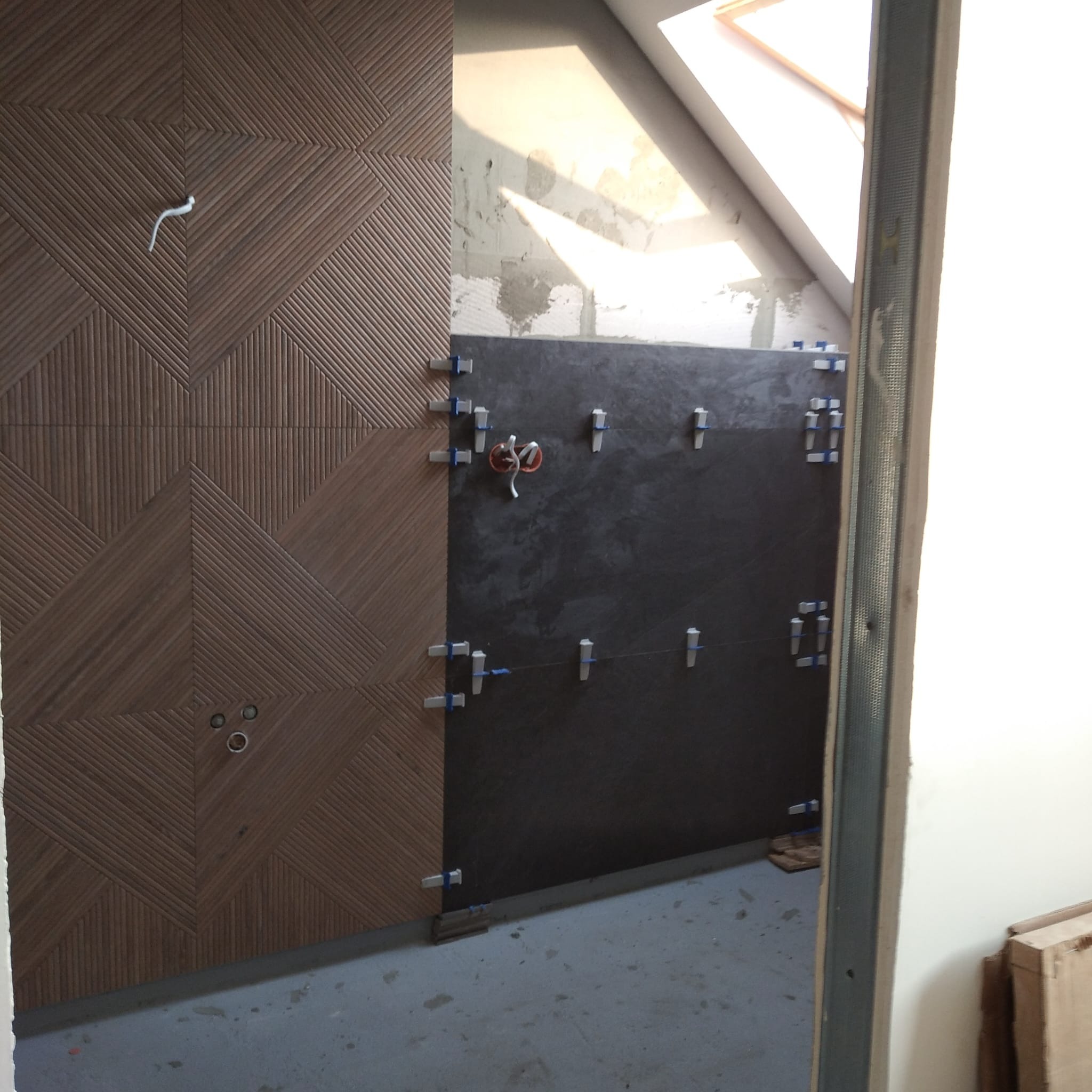 Układanie dekoracyjnych płytek w łazience na poddaszu w Poznaniu. Widoczne elementy montażowe i fragment surowej ściany. Nietypowy wzór płytek.