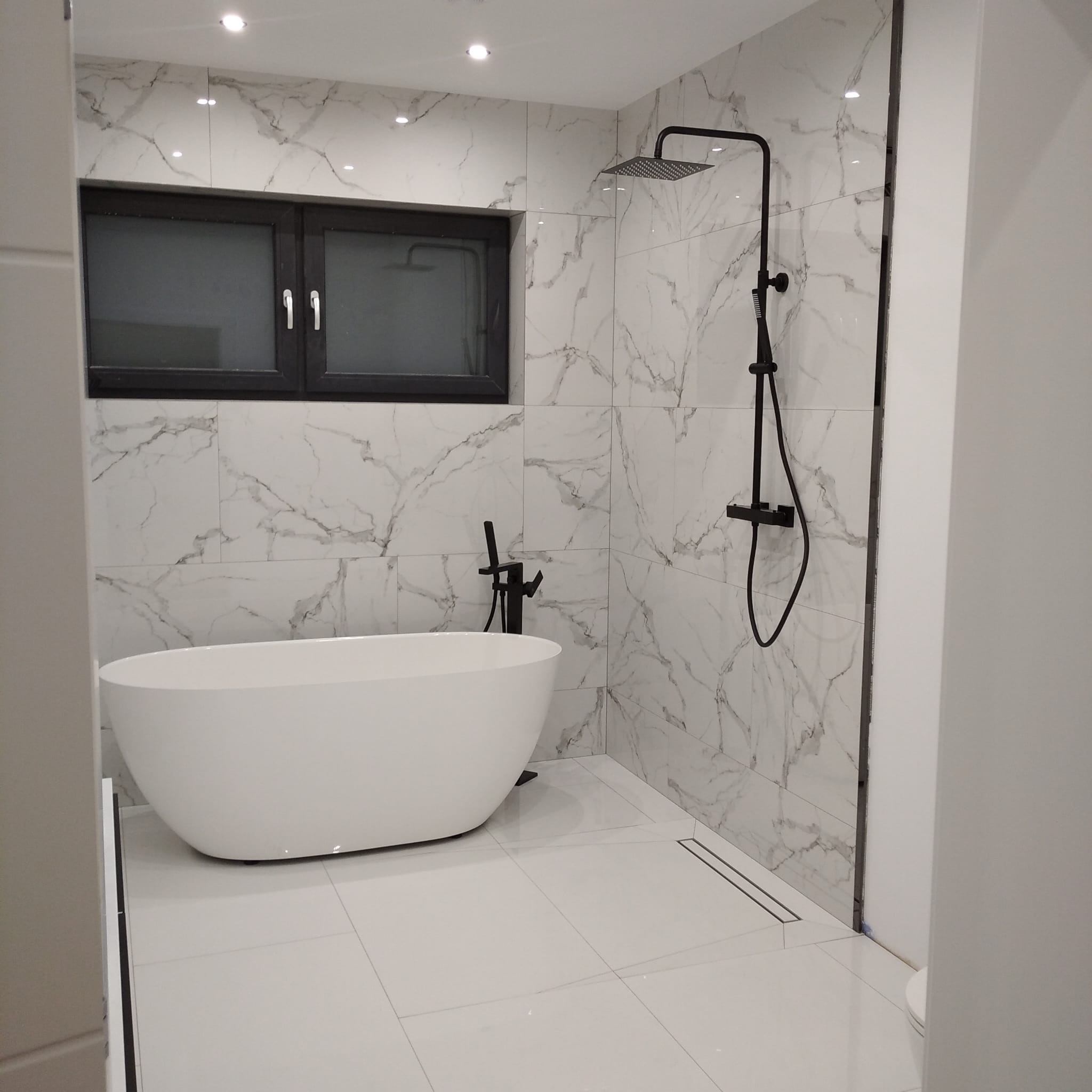 Nowoczesna łazienka z białą wanną wolnostojącą, czarną armaturą i płytkami imitującymi marmur. Minimalistyczny design z oknem i odpływem liniowym.