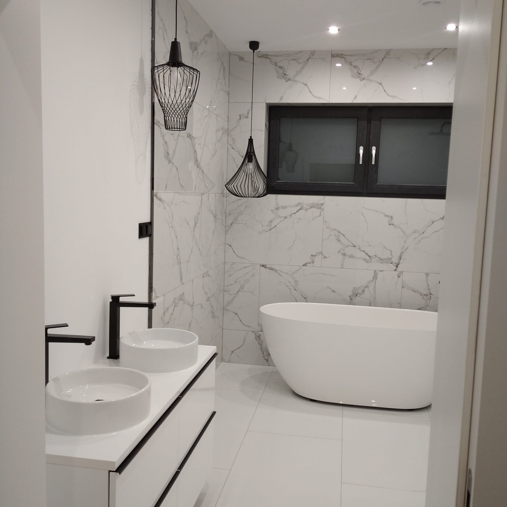 Nowoczesna łazienka z marmurowymi płytkami, wolnostojącą wanną i podwójną umywalką na szafce z czarnymi akcentami. Dwie wiszące lampy o ażurowej konstrukcji.